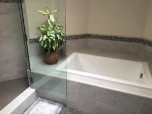 This image features a bathtub with grey tile as well as a plant resting next to the end of the tub. 